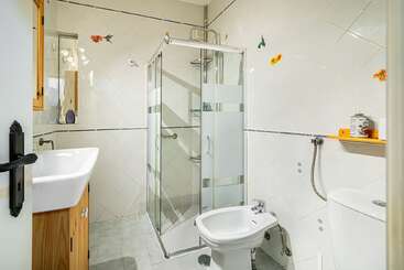 This bathroom features a glass shower enclosure, white tiles with decorative stickers, a bidet, toilet, wooden vanity with sink, wall shelf, and ample lighting.