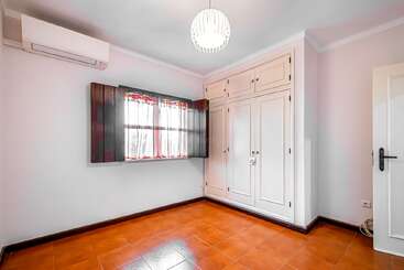 This image shows an empty room with orange tile flooring, white walls, a large built-in wardrobe, a window with shutters, an air conditioner, and overhead lighting.