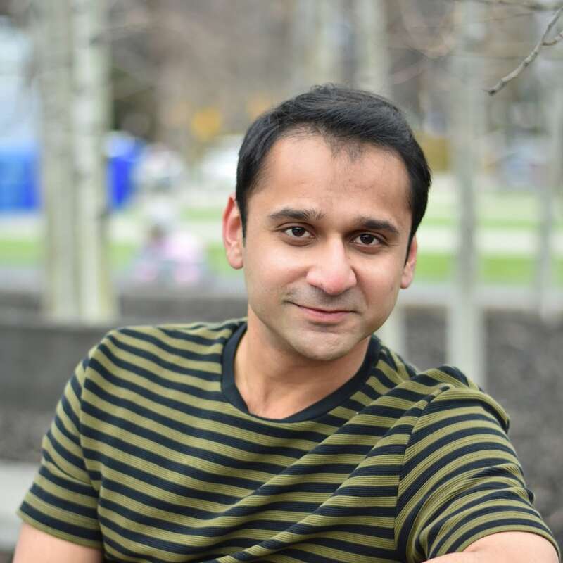 A man with short dark hair is wearing a green and black striped t-shirt, smiling slightly, seated outdoors with blurred trees and park in the background.