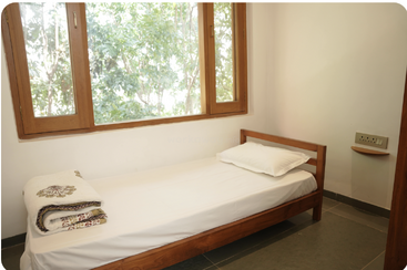 This image shows a simple, clean bedroom with a single wooden bed, white linens, large windows, folded blankets, and a view of green trees outside.
