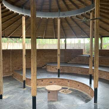 This image shows a circular, open-roofed amphitheater made of natural materials, with tiered seating, bamboo pillars, stone floor, and a central round platform. Natural light enters.