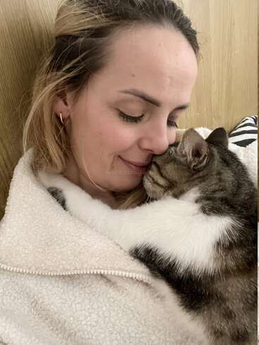 Una mujer abraza cariñosamente a su gato, ambos con los ojos cerrados. El gato se acurruca estrechamente, envolviéndola con sus patas, creando un cálido y tierno momento de afecto.