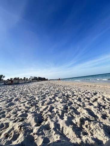Amplia playa de arena bajo un cielo azul vibrante, con suaves olas marinas. La gente camina por la orilla, palmeras y edificios lejanos se perfilan en el horizonte. Ambiente tranquilo.