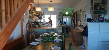 Une cuisine et une salle à manger confortables avec un escalier en bois, une table dressée, deux personnes faisant la vaisselle, des étagères remplies de livres et un éclairage chaleureux créant une atmosphère accueillante.