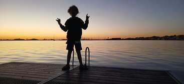Un enfant se tient debout sur un quai au coucher du soleil, se découpant en silhouette sur un lac calme. L'eau paisible, les bateaux au loin et le ciel orange pâle créent une scène sereine.