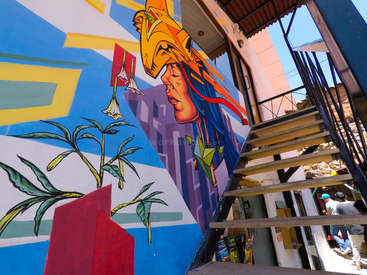 A imagem mostra um mural vibrante no exterior de um edifício, com o rosto de uma mulher e uma planta, com uma escada que leva a uma entrada e pessoas caminhando nas proximidades.