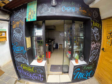 A imagem mostra a fachada de um restaurante com uma parede externa preta com arte colorida em giz e uma porta de vidro, mostrando o interior da cozinha.
