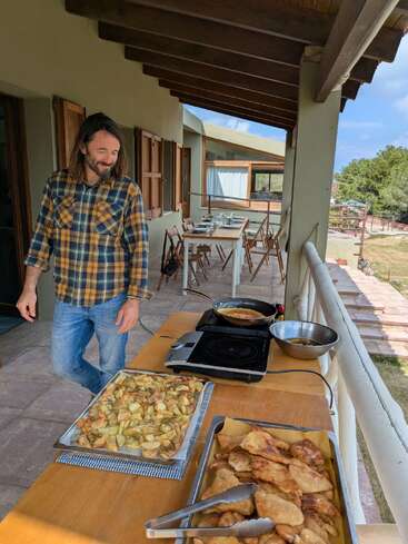 Un hombre con una camisa a cuadros está de pie en un porche bañado por el sol, sonriendo. Cerca de allí, hay bandejas con patatas cocidas y comida frita preparadas para la comida.