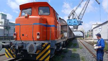 Une locomotive orange est garée sur les rails près d'un site industriel. Un ouvrier vêtu de bleu observe la scène tandis qu'une grande grue opère au-dessus de lui sous un ciel partiellement nuageux.