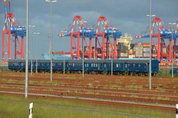 Au premier plan, un train de voyageurs bleu est immobilisé sur les rails. À l'arrière-plan, des grues colorées et des conteneurs empilés dominent le port industriel en pleine effervescence.