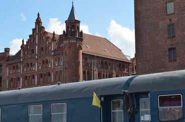 Un bâtiment historique en briques rouges, à l'architecture richement décorée et surmonté d'une tour pointue, se dresse derrière un wagon de train bleu foncé, sous un ciel bleu vif parsemé de nuages.