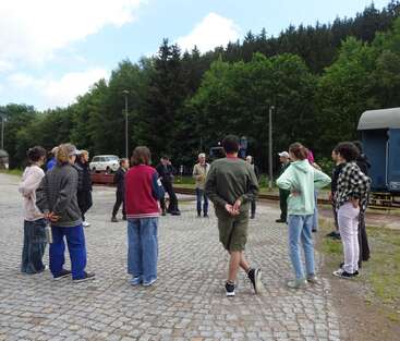 Un groupe de personnes est debout en cercle sur une esplanade pavée, près d'un wagon bleu portant l'inscription « DAS LETZTE KLEINOD », au milieu des arbres.