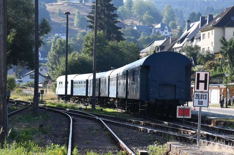 L'image représente un train sur une voie ferrée, avec une rangée de maisons à l'arrière-plan et une zone herbeuse au premier plan.