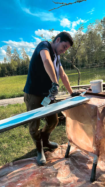 Ein Mann ist an einem sonnigen Tag im Freien und streicht eine Holzplanke blau an. Er trägt Handschuhe und Stiefel und arbeitet auf einer provisorischen Fläche, die von Bäumen umgeben ist.