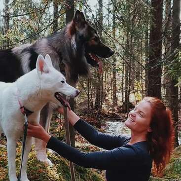 A woman with red hair smiles joyfully at two large dogs, one white and one black, as they enjoy a walk together in a lush forest.