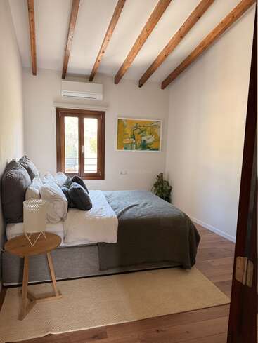 Dieses gemütliche Schlafzimmer verfügt über Holzbalken an der Decke, ein großes Bett mit grau-weißer Bettwäsche, ein abstraktes Kunstwerk, Holzböden und einen kleinen runden Tisch.
