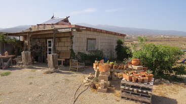 A rustic stone house stands in an arid landscape, surrounded by potted plants, wooden furniture, and greenery. Mountains rise in the hazy distance behind it.