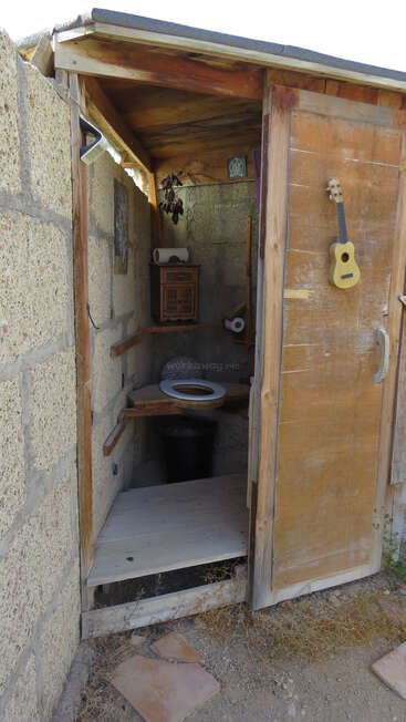 This image shows a rustic outdoor outhouse with a wooden door, stone walls, composting toilet, paper rolls, and a small ukulele hanging on the door.