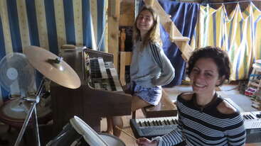 The image shows two women in a room with musical instruments, smiling at the camera, surrounded by a staircase and colorful curtains.