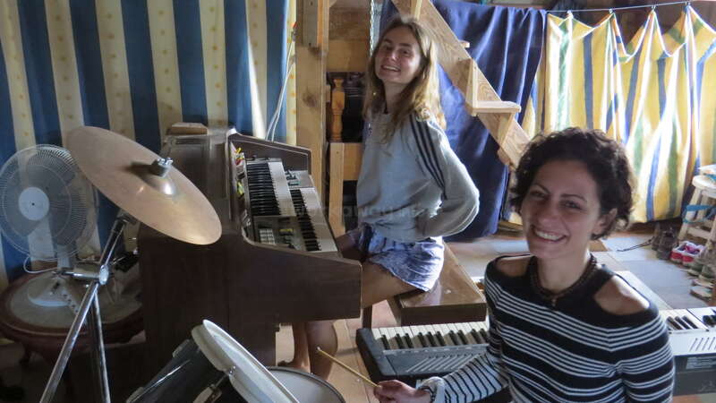 The image shows two women in a room with musical instruments, smiling at the camera, surrounded by a staircase and colorful curtains.