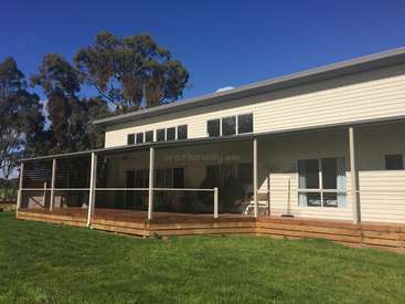 The image depicts a modern, single-story house with a spacious wooden deck and a lush green lawn, set against a clear blue sky.