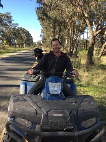 The image shows a man riding an ATV on a road, with a dog sitting behind him, surrounded by trees and grass.