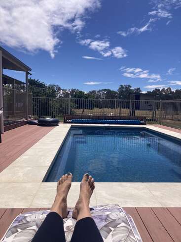 A person relaxes on a lounge chair by an inviting blue swimming pool, under a partly cloudy sky, surrounded by a fence and trees in the background.