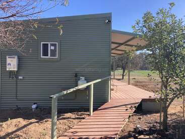 A small green corrugated metal building with a wooden ramp and covered porch sits amidst trees and open land, under a clear blue sky.