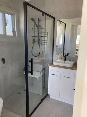 This modern bathroom features a glass shower with black trim, a handheld showerhead, a shower chair, a white vanity with a vessel sink, and grey tile walls.