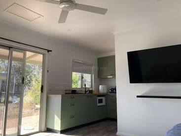 This image shows a modern kitchen with green cabinets, wooden countertops, and appliances. There’s a large TV on the wall and sliding glass doors to the outside.