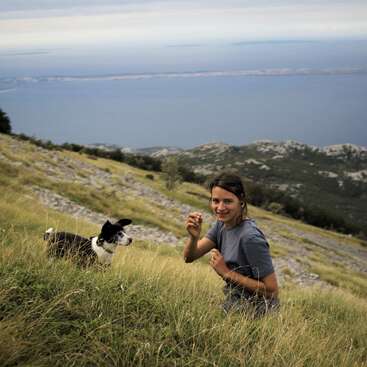 A woman and her black-and-white dog are on a grassy hillside with scenic mountains and a blue sea in the background, enjoying a peaceful outdoor adventure.