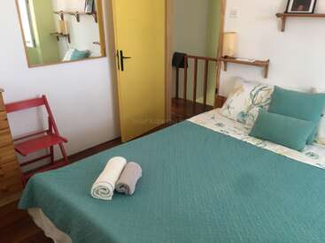 This cozy bedroom features a teal bedspread, yellow door, red chair, wooden shelves, and a mirror. Towels are neatly placed on the bed, creating welcoming vibes.