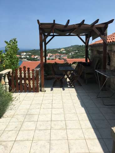 A tiled terrace with a shaded wooden pergola overlooks a coastal village. Outdoor furniture and a drying rack complete this sunny, relaxing scenic spot.