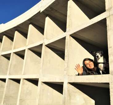 A woman and her dog are inside a concrete grid structure, smiling and waving. Sunlight casts sharp shadows, highlighting the geometric architecture and creating a cheerful scene.