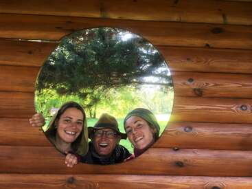 Three smiling people, two women and one man, peek joyfully through a large round window in a wooden wall, with green trees and sunlight behind them.