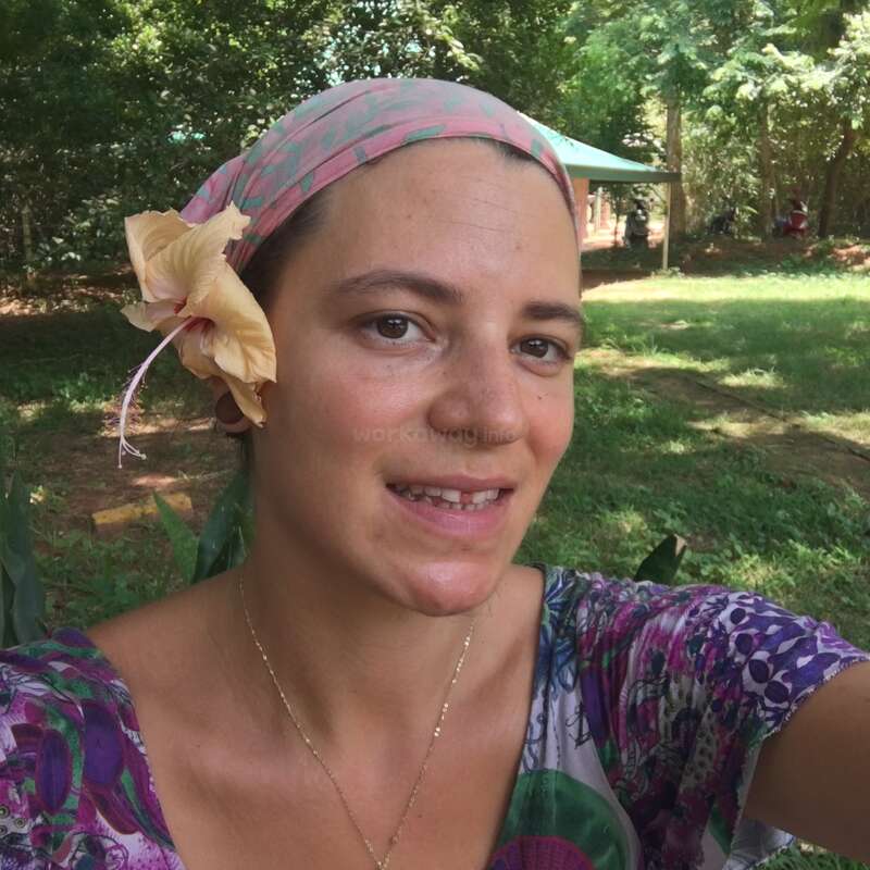 A woman takes a selfie outdoors, wearing a colorful headscarf and a hibiscus flower in her hair. She smiles slightly, surrounded by greenery and trees.