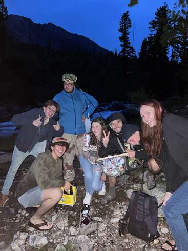 Six friends pose together outdoors in the evening, surrounded by trees and mountains. They smile, hold drinks, and appear to be enjoying a fun gathering.