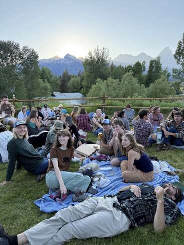 Un grupo de personas disfruta de un pícnic en un prado al atardecer, rodeados de árboles y montañas, compartiendo comida, bebida y risas sobre unas mantas.