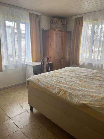 Dieses gemütliche Schlafzimmer hat ein großes Bett, einen Kleiderschrank aus Holz, einen Eckschreibtisch mit Stuhl, Fliesenboden und zwei helle Fenster mit durchsichtigen und gelben Vorhängen.