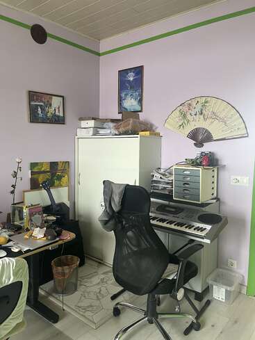Ein Homeoffice mit Tastatur, schwarzem Stuhl, vollgestopftem Schreibtisch, Schrank, Wanddeko, Deko-Ventilator, Papierkram und pastellfarbenen lila Wänden mit grünen Akzenten. Gemütliche Atmosphäre.