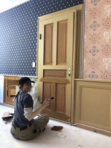 A woman sits on the floor, smiling while painting a wooden door. The room features patterned wallpaper and painted paneling, with painting supplies scattered around her.