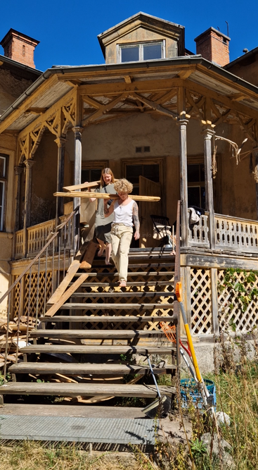 Two people descend wooden stairs of an old, rustic house under renovation, carrying wood planks. The weather is sunny, with clear blue skies and visible tools.