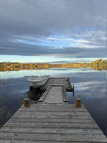 A wooden dock leads to a calm lake, with a small rowboat tied at the end. Autumn trees reflect on the serene, still water.