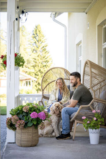 A smiling couple relaxes on a cozy porch with their fluffy dog, surrounded by blooming flowers in baskets and pots. Outdoor furniture creates a peaceful atmosphere.