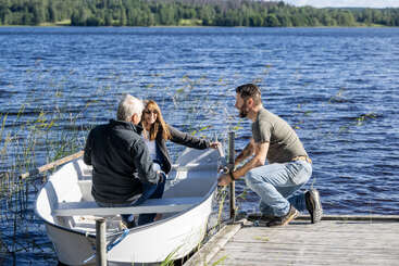 Three people enjoy a sunny day by the lake; two sit in a small white boat, while another kneels on the dock, helping them prepare to depart.