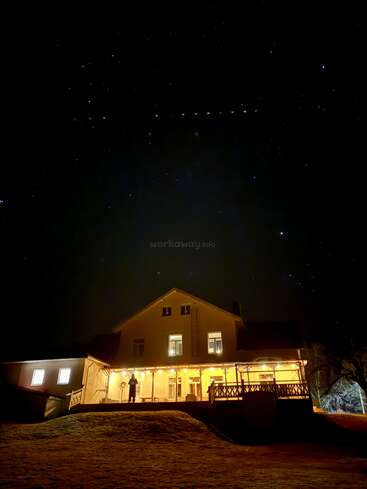 A warmly lit house shines against a dark, star-filled night sky. One person stands on the porch. Shadows and soft snow surround the peaceful, inviting scene.