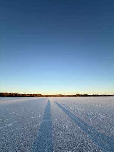A frozen lake stretches to the horizon, sparkling under clear blue skies. Two long shadows of people are cast across the icy surface at sunset.