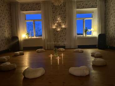 A cozy meditation room with round cushions arranged in a circle, lit candles, soft lighting, floral wallpaper, large windows, and calming evening ambiance. Peaceful and inviting atmosphere.