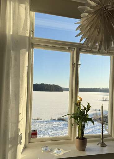 Sunlight streams through a window overlooking a snowy landscape and distant trees. On the windowsill sits a potted plant, candleholder, and two glass candle holders.