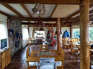 The image depicts a spacious room with wooden beams, a stone fireplace, and a Christmas tree, featuring a dining table, chairs, and a couch, with a TV on a cabinet to the left.